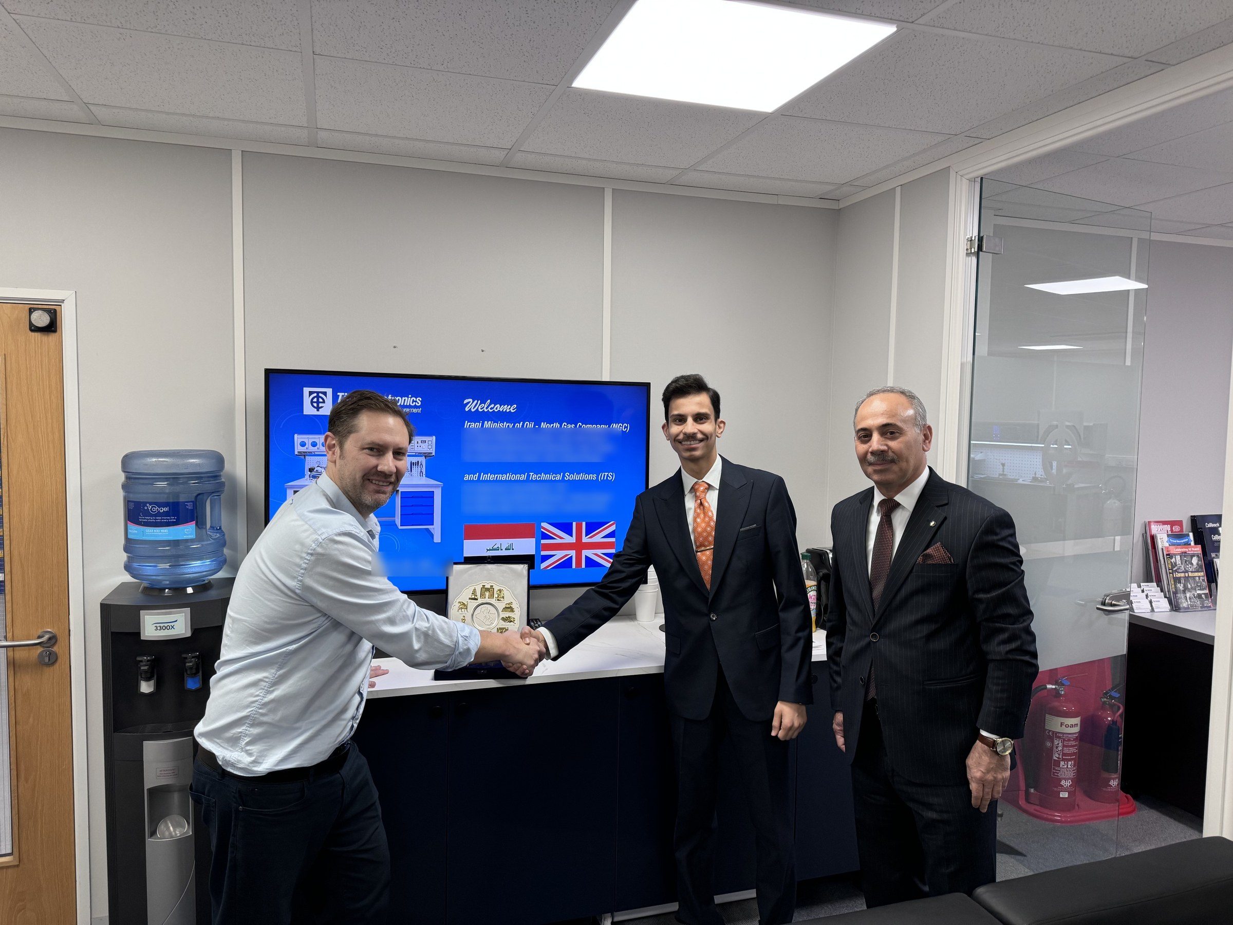 Robert Bailey of Time Electronics shaking hands with Omar Farooq Al Obaidi under a welcome screen showing the Iraqi and British flags.