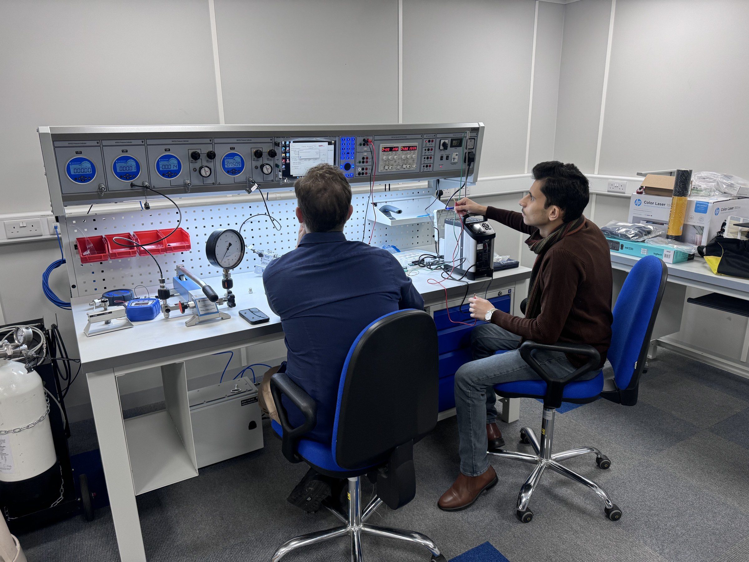 Omar Farooq Al Obaidi working the instruments of a calibration bench beside a Time Electronics engineer.